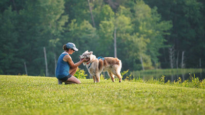 Woman bends over to pet her senior dog in the grass by a pond.